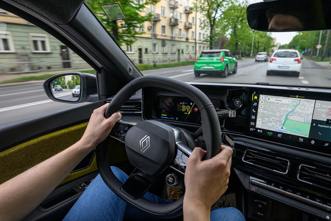 Widok zza kierownicy samochodu, na ulicy z pasem zieleni i kilkoma budynkami mieszkalnymi. Kierowca trzyma kierownicę, na panelu widoczna jest nawigacja GPS pokazująca trasę. Przed pojazdem widać inne samochody na drodze.