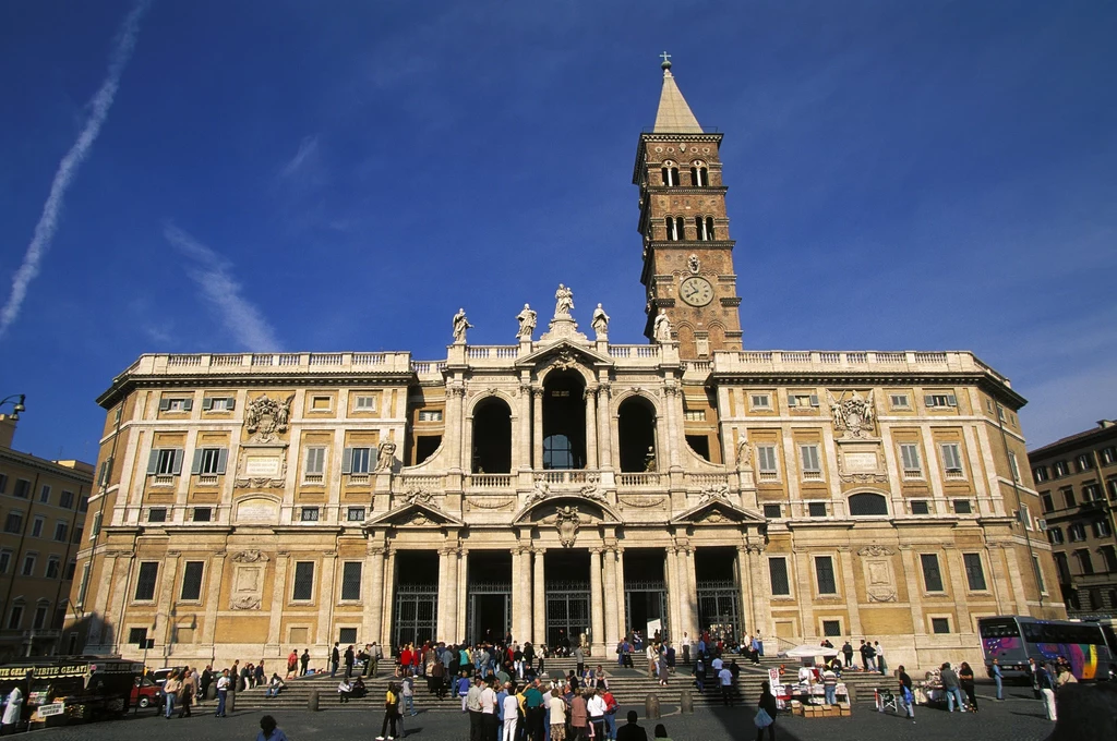 Bazylika Matki Bożej Śnieżnej Bazylika Santa Maria Maggiore w Rzymie z charakterystyczną fasadą i wysoką dzwonnicą, wokół której gromadzą się ludzie. Budynek wyróżnia się bogatymi detalami architektonicznymi i historyczną fasadą.