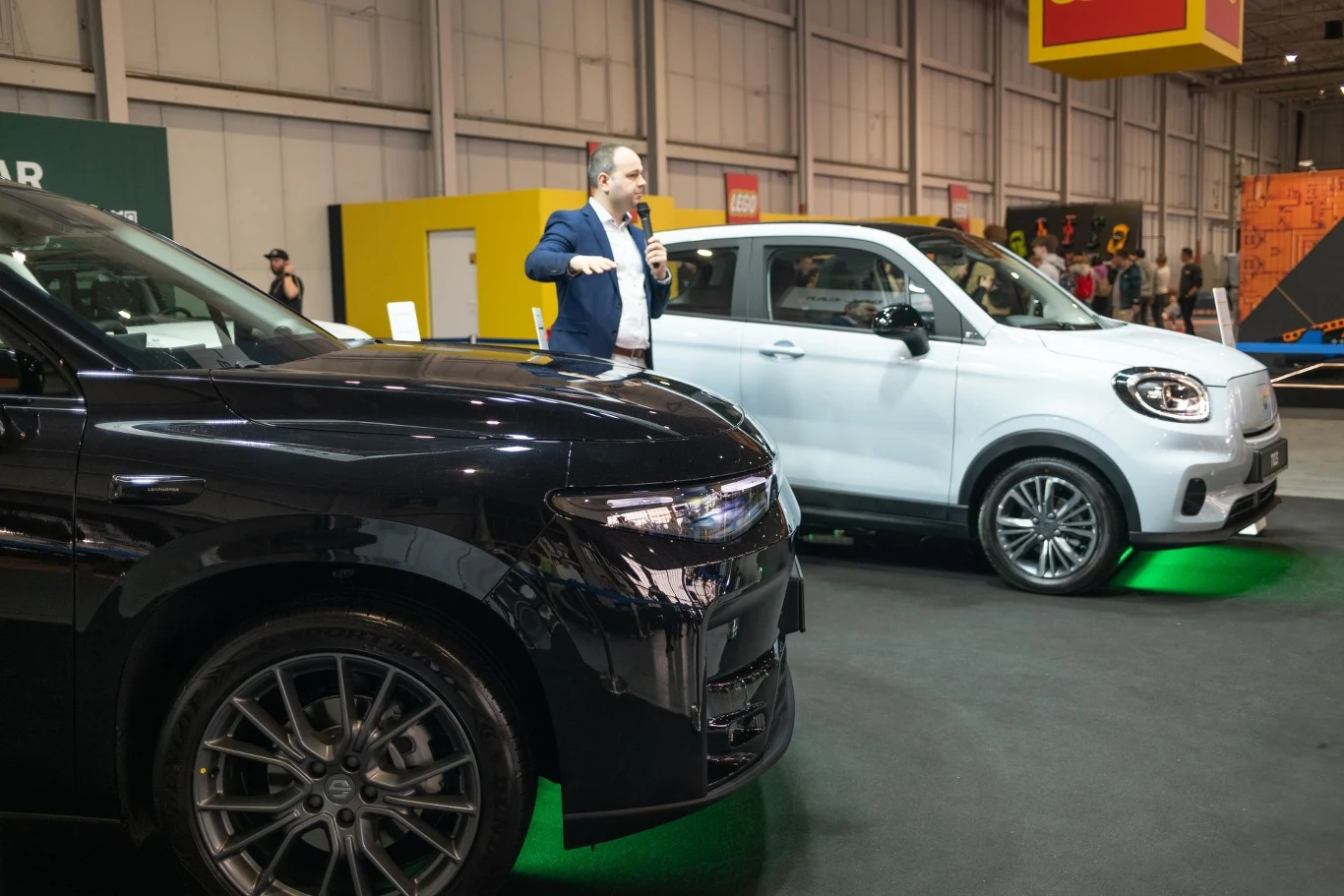 Leapmotor obecna na targach Poznań Motor Show 2025. Dwa samochody wystawione na targach motoryzacyjnych, jeden czarny i drugi biały, z mężczyzną z mikrofonem, który prawdopodobnie prowadzi prezentację. W tle widoczne są stoiska wystawowe.