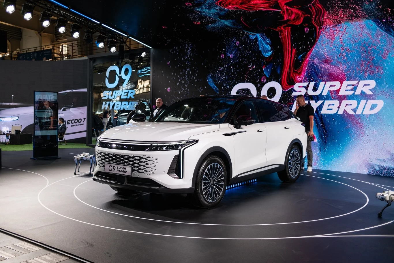 Ekskluzywny samochód super hybrydowy w kolorze białym prezentowany na wystawie motoryzacyjnej. Otoczony przez dynamiczne tło z logo i efektami świetlnymi, pojazd przyciąga uwagę jako centralny element ekspozycji.