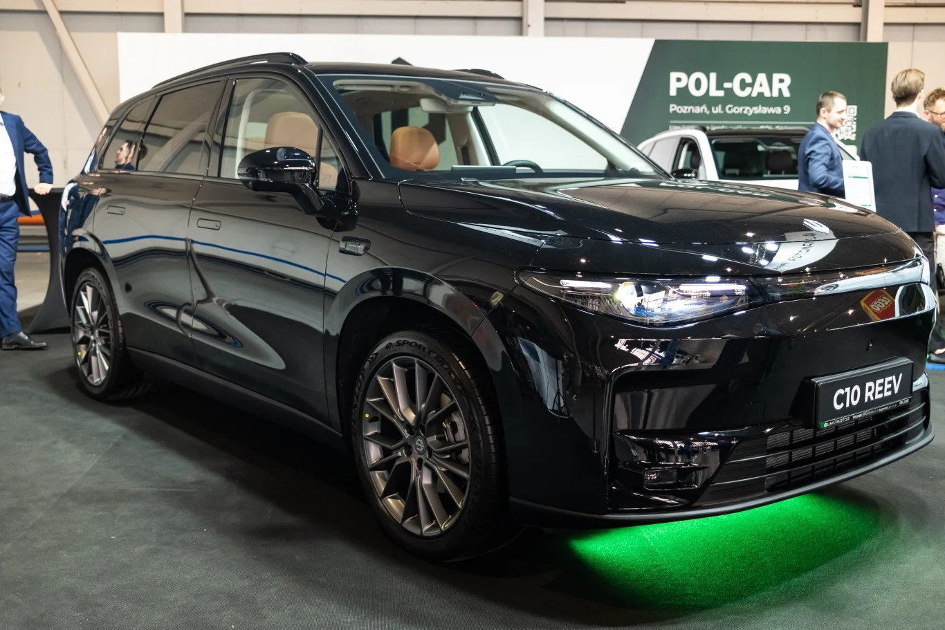 Nowością zaprezentowaną na targach jest Leapmotor C10 REEV Czarny samochód o eleganckim wyglądzie, podświetlenie podwozia zielonym światłem, stoi na wystawie w pomieszczeniu, w tle widoczne osoby i banner reklamowy.