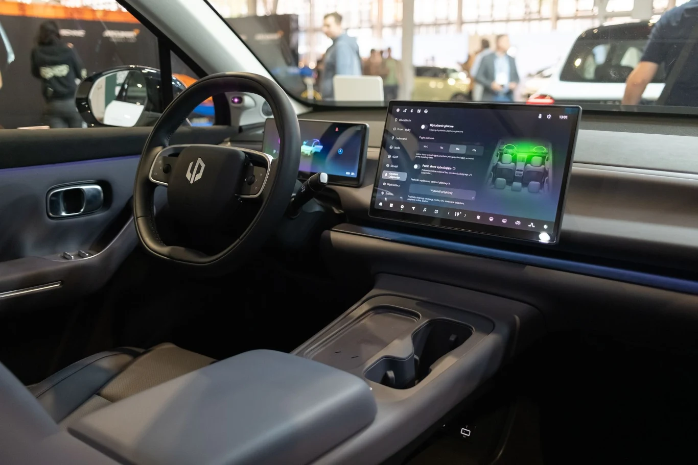 Przyszłość marki Leapmotor w Polsce. Powstają nowe punkty sprzedaży i serwisu. Wnętrze nowoczesnego samochodu elektrycznego z dużym wyświetlaczem dotykowym na desce rozdzielczej. Kierownica wraz z cyfrowym panelem instrumentów. W tle widoczne postacie rozmawiające na targach motoryzacyjnych.
