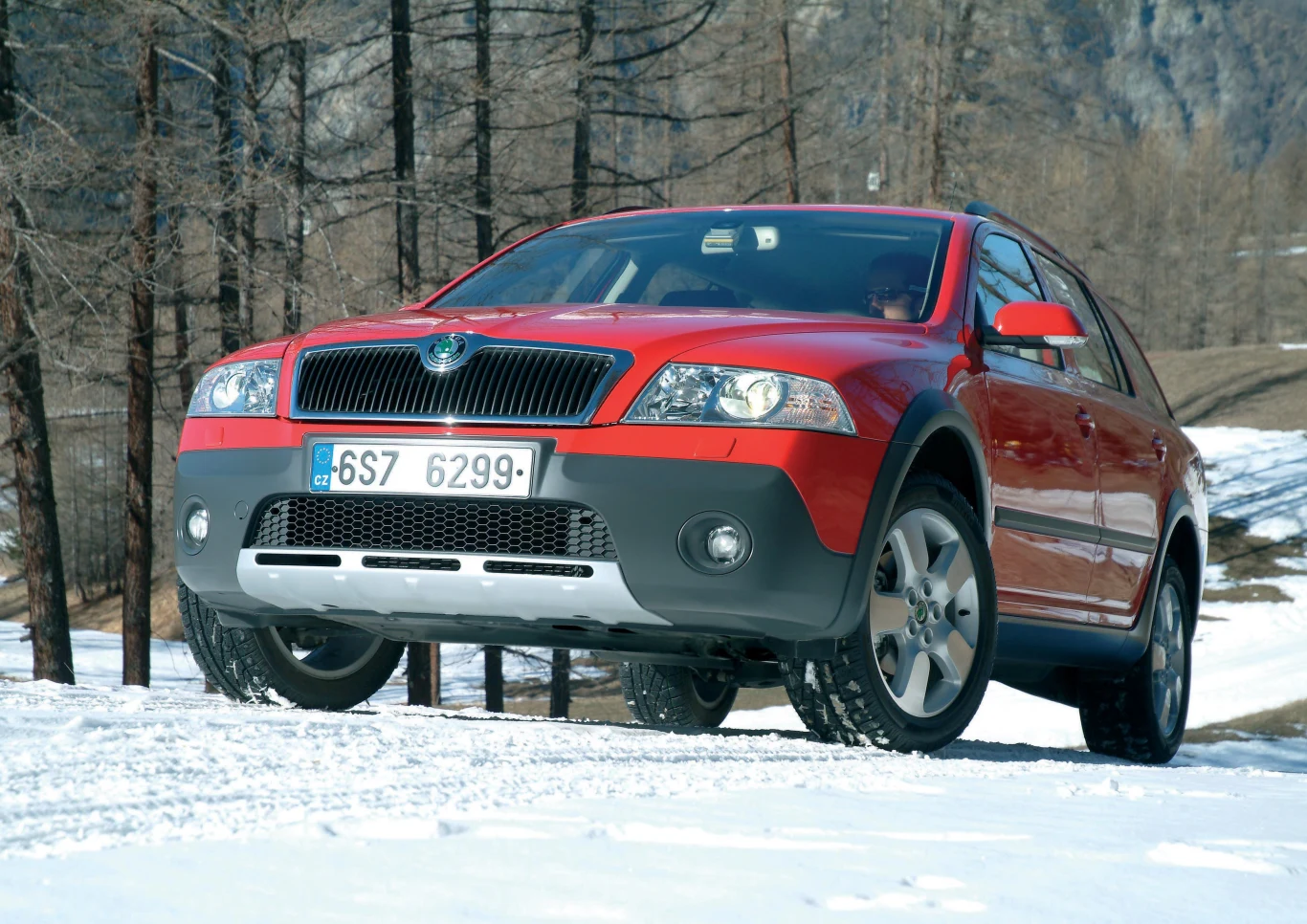 Czerwony SUV na zaśnieżonej drodze w lesie. Samochód ma szerokie opony, a wokół znajdują się nagie drzewa, co sugeruje zimową porę roku. Tło stanowią góry, widać dynamiczny kadr z pojazdem na pierwszym planie.