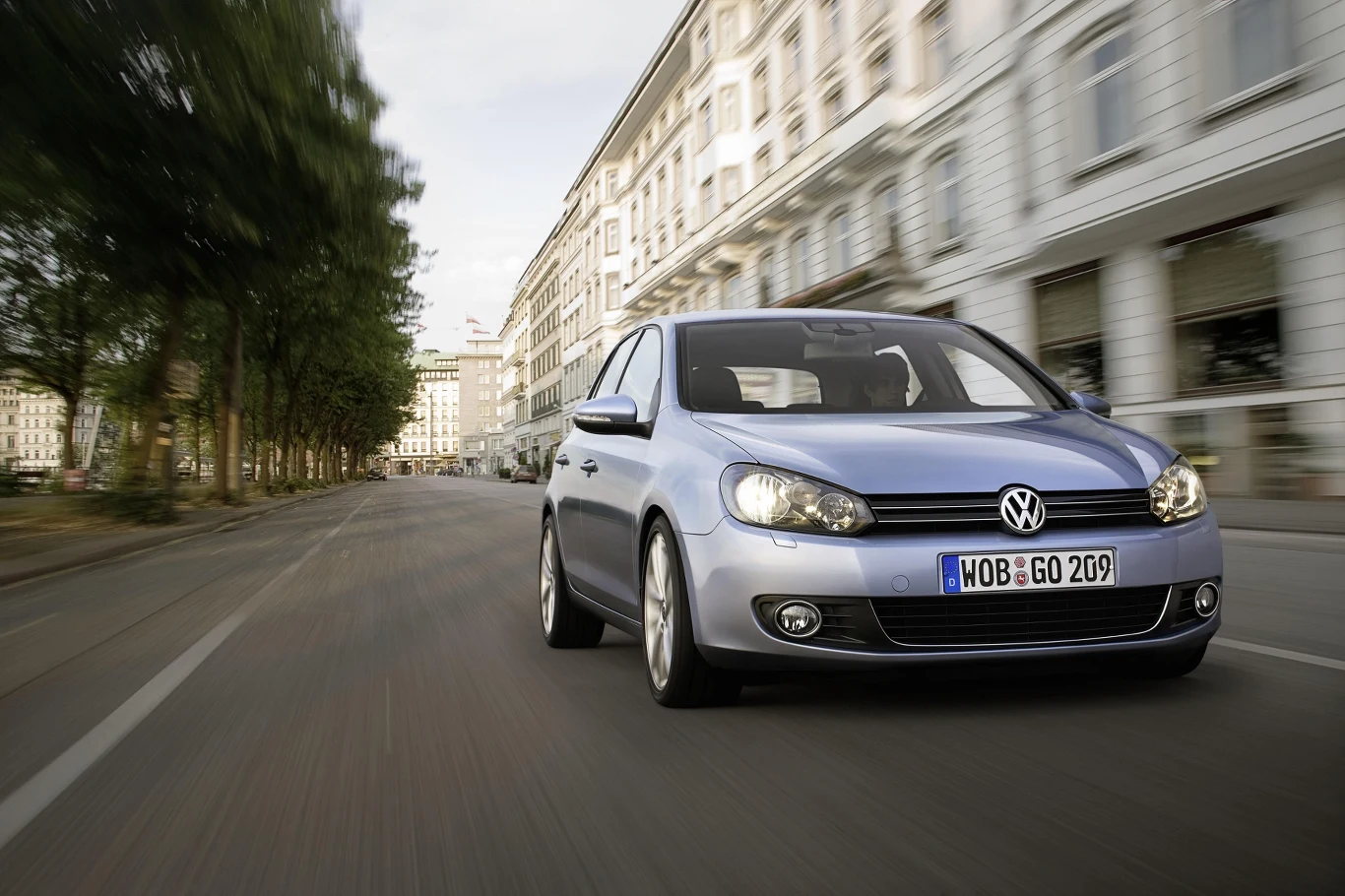 Srebrny samochód Volkswagen porusza się dynamicznie po szerokiej, pustej ulicy otoczonej wysokimi, eleganckimi budynkami.