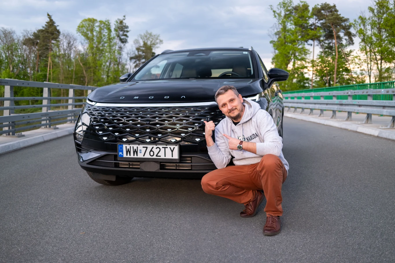 Omoda 5 to moim zdaniem najlepsze spalinowe auto z Chin, jakie można dziś kupić w Polsce / fot. Piotr Król Mężczyzna siedzący przy czarnym SUV-ie z rejestrem WW-762TY na moście. W tle drzewa i niebo.