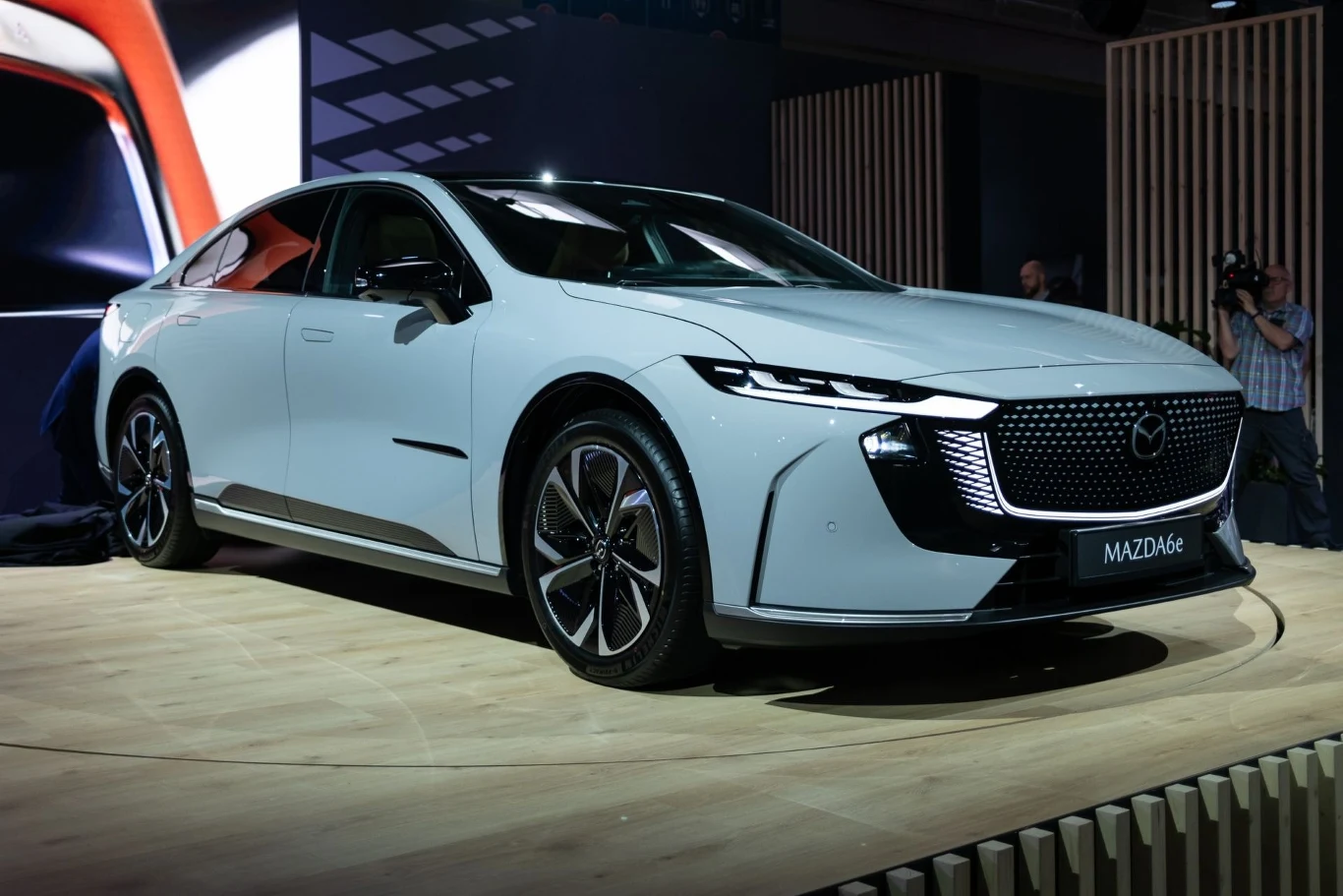 Mazda 6e oficjalnie zadebiutowała w Polsce. Ceny robią wrażenie./fot. Piotr Król Biały samochód osobowy marki Mazda, o nowoczesnym i eleganckim designie, stoi na drewnianej platformie wystawowej na targach motoryzacyjnych. W tle widać ludzi robiących zdjęcia oraz ekran z fragmentem auta w dużym zbliżeniu.