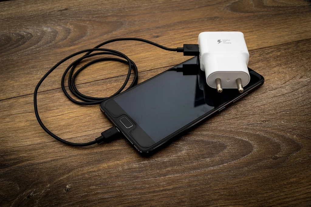 Fot. pixabay.com/ ReadyElements Smartfon z czarnym ekranem leży na drewnianej powierzchni, obok niego znajduje się biała ładowarka z kablem USB. Wszystko jest ułożone w sposób schludny i widoczne w jasnym świetle.