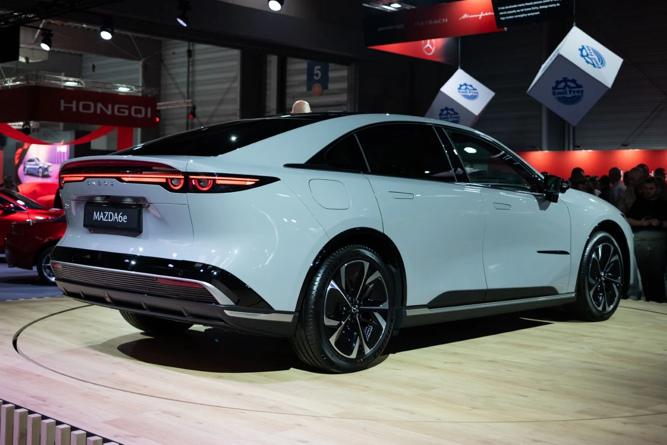 Mazda 6e dostępna jest w dwóch wariantach napędu: 258 KM i akumulator 68,8 kWh oraz 244 KM i 80 kWh./fot. Piotr Król Targowa prezentacja nowoczesnego, białego samochodu elektrycznego o nazwie MAZD6e. W tle widoczne są inne stoiska oraz elementy wystroju targów motoryzacyjnych.