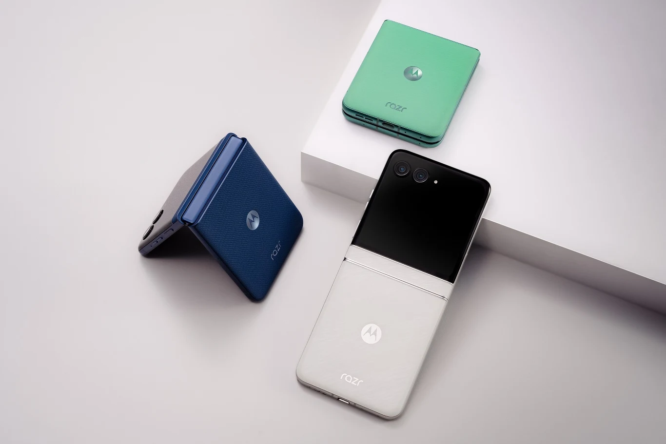 Trzy nowoczesne smartfony typu flip na białym tle, każdy w innym kolorze: niebieskim, zielonym i czarnym, połączonym z białym. Telefony są ustawione w różnych pozycjach, co pokazuje ich konstrukcję i zamknięty stan.