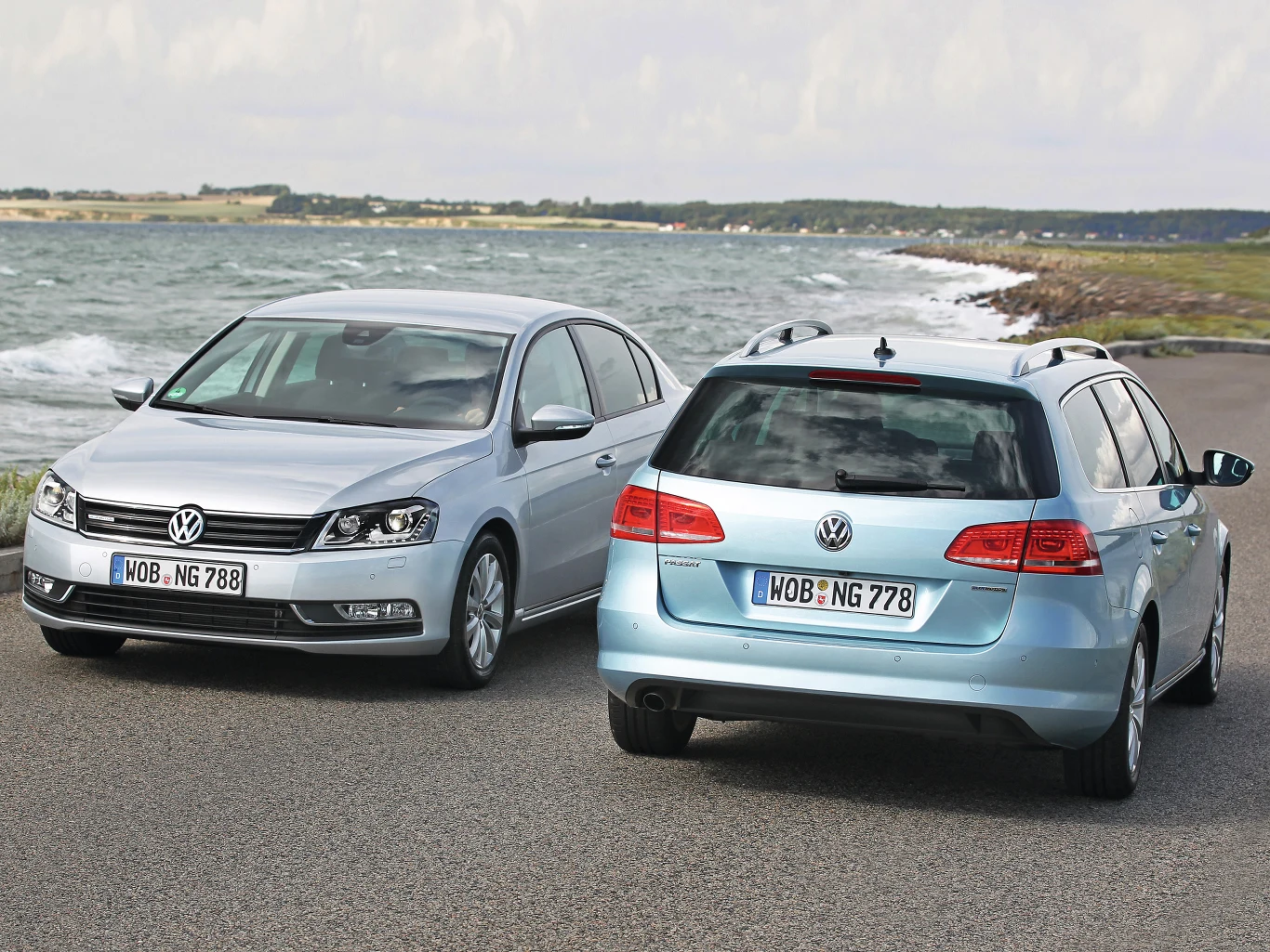Dwa samochody Volkswagen nad brzegiem morza, jeden widoczny z przodu, drugi z tyłu. Oba pojazdy stoją na asfaltowej drodze, w tle widać linię brzegową i lekko zachmurzone niebo.