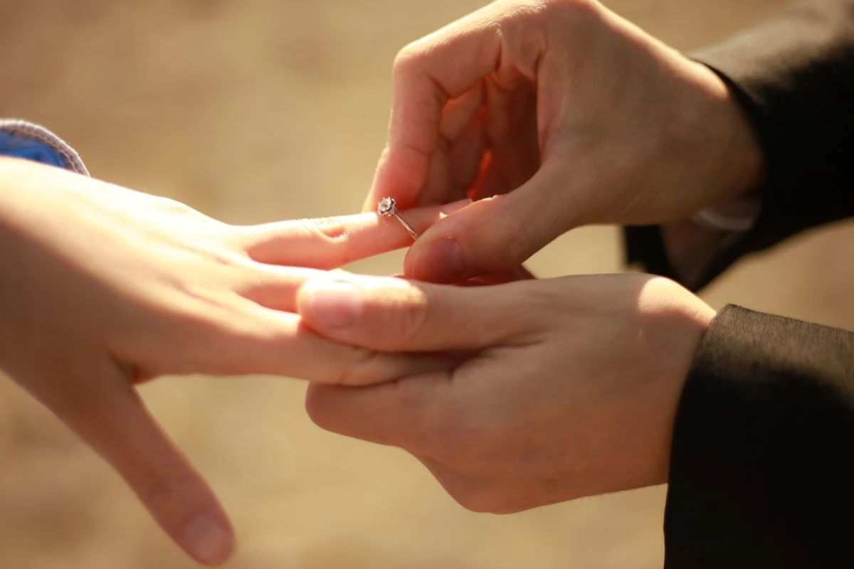 Mężczyzna wkłada pierścionek na palec kobiety, co sugeruje moment zaręczyn. Dłonie są delikatnie oświetlone i widać na nich emocje związane z tą wyjątkową chwilą.