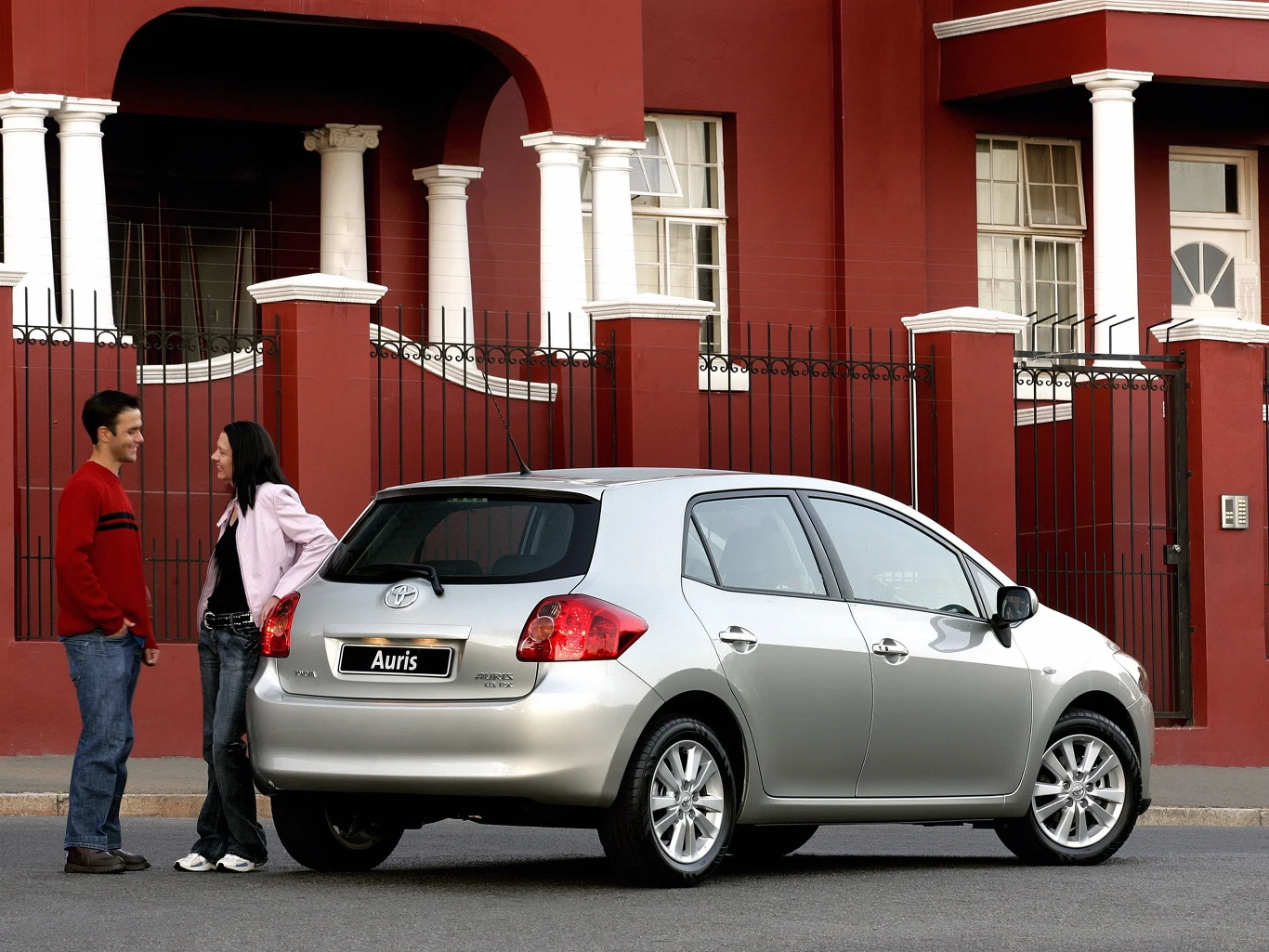 Auris I ma też problemy z korozją, ale mimo wszystko mniejsze niż Civic Srebrny samochód Toyota Auris zaparkowany przed eleganckim, czerwonym budynkiem z kolumnami. Obok auta stoją mężczyzna i kobieta rozmawiający ze sobą. Scena wygląda na przyjacielską i spokojną.