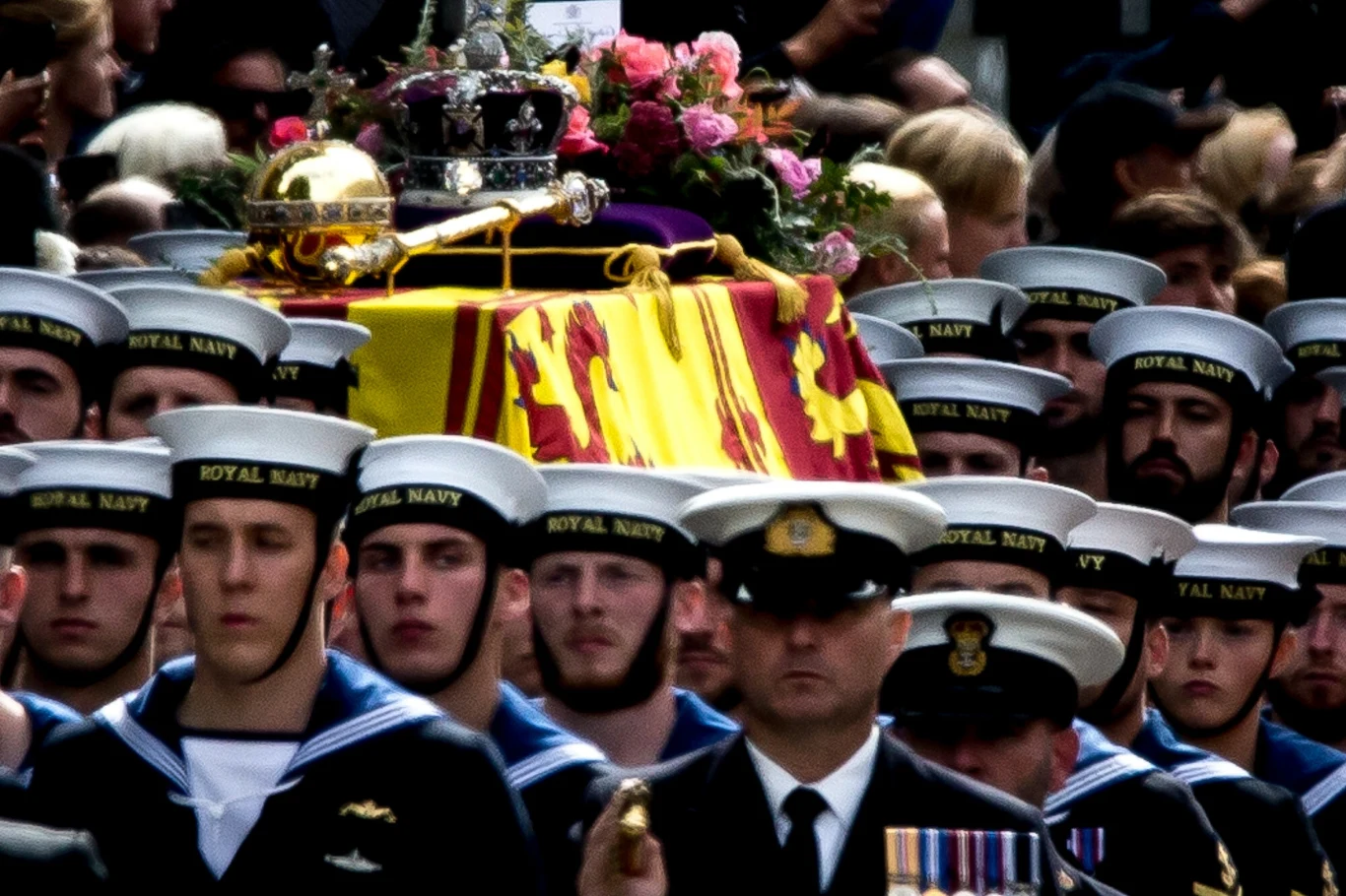 Procesja pogrzebowa królowej Elżbiety II Żołnierze Królewskiej Marynarki Wojennej w mundurach, niosą trumnę ozdobioną koroną i flagą, otoczeni uczestnikami uroczystości. Trumna jest dodatkowo ozdobiona kolorowymi kwiatami.