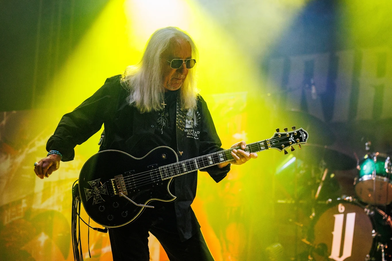 Mick Box jest liderem Uriah Heep Muzyk z długimi, siwymi włosami gra na czarnej gitarze elektrycznej na scenie, oświetlony żółtym i zielonym światłem. W tle widoczne są perkusja i efekty świetlne, tworzące intensywną atmosferę koncertową.