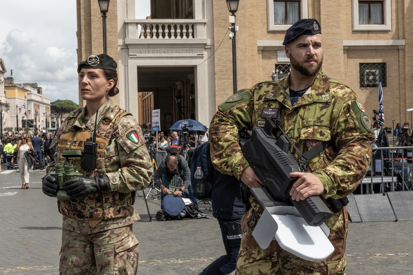 Włoscy żołnierze patrolujący tereny Placu Świętego Piotra z bronią antydronową Dwoje żołnierzy w mundurach armii włoskiej stoi na placu w mieście. Kobieta trzyma lornetkę, a mężczyzna nosi broń. W tle widoczni są ludzie i budynki miejskie.