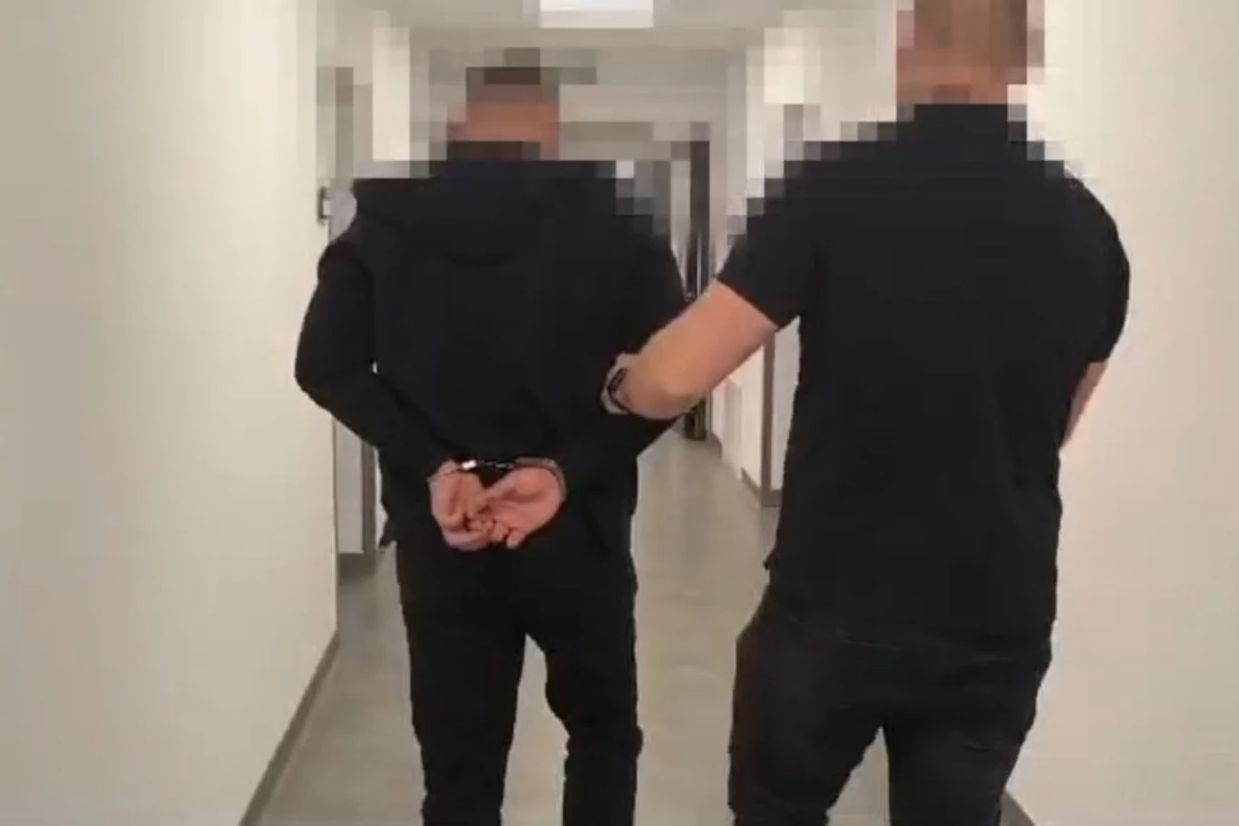 Dwóch mężczyzn w korytarzu, jeden prowadzi drugiego skuta kajdankami, a ich twarze są rozmyte dla ochrony tożsamości.