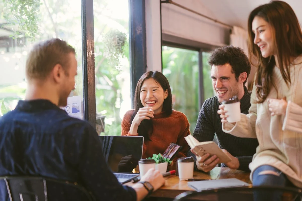 Grupa młodych ludzi siedzi przy stole w kawiarni, uśmiechając się i rozmawiając. Na stole widać laptopa i książki, co może sugerować luźne spotkanie towarzyskie lub sesję roboczą.