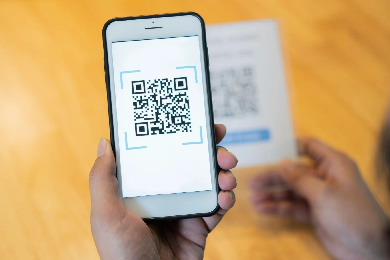 Uważaj na podejrzane kody QR przy twoim samochodzie. Ręka trzymająca smartfon skanujący kod QR, w tle niewyraźna karta z innym kodem. Styl minimalistyczny, drewniane tło dodaje ciepła scenie, główny punkt to wyświetlany kod na ekranie telefonu.