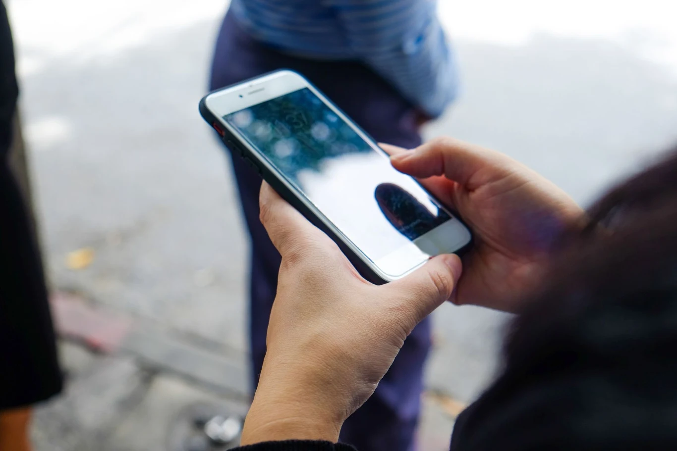 Zeskanowany fałszywy kod QR może przysporzyć wielu kłopotów. Osoba trzymająca w ręku smartfon, patrząca na jego ekran. W tle widoczna jest nieostro zarysowana postać oraz fragment chodnika.