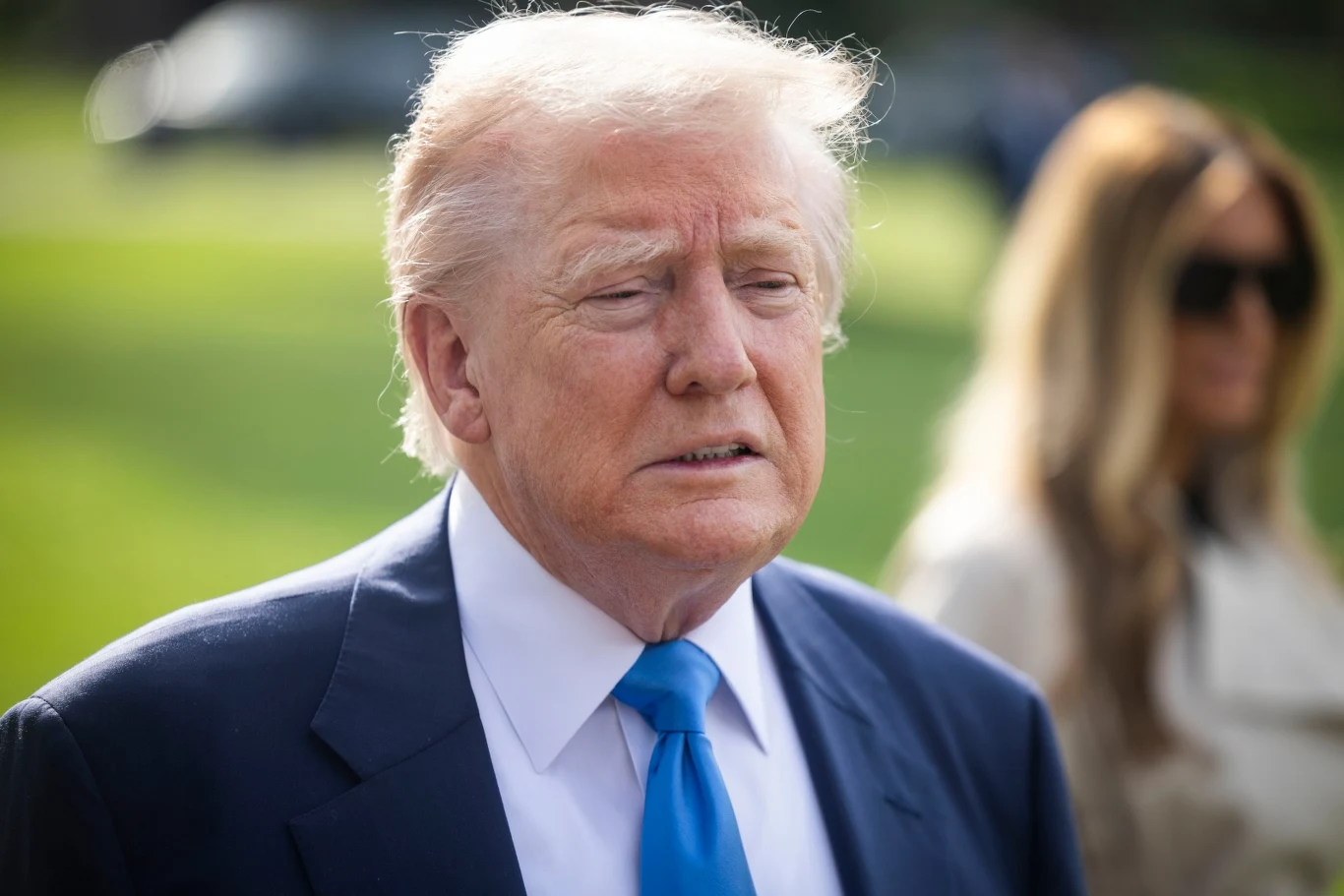 Donald Trump zabrał głos ws. umowy o rzadkich minerałach Mężczyzna w ciemnym garniturze i niebieskim krawacie stoi na zewnątrz. W tle niewyraźna postać kobiety. Obszar zieleni wskazuje na plener.