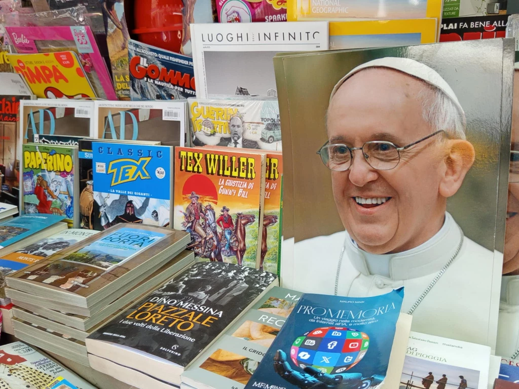 Papież "spoglądający" na komiksy stoisko z różnorodnymi czasopismami i książkami, na pierwszym planie duży plakat z wizerunkiem papieża, obok wiele publikacji o tematyce religijnej, kulturalnej i rozrywkowej