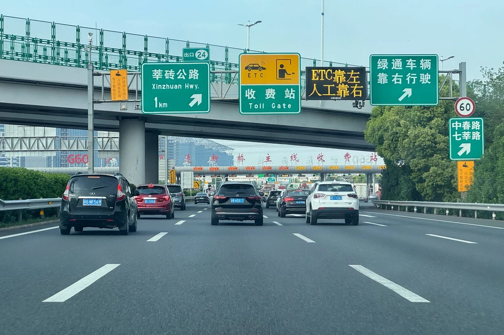 Tablice informacyjne nad drogami w Chinach mają angielskie tłumaczenie Autostrada z kilkoma pasami ruchu wypełnionymi samochodami. W tle znajdują się znaki drogowe wskazujące kierunki jazdy oraz konstrukcja mostu.