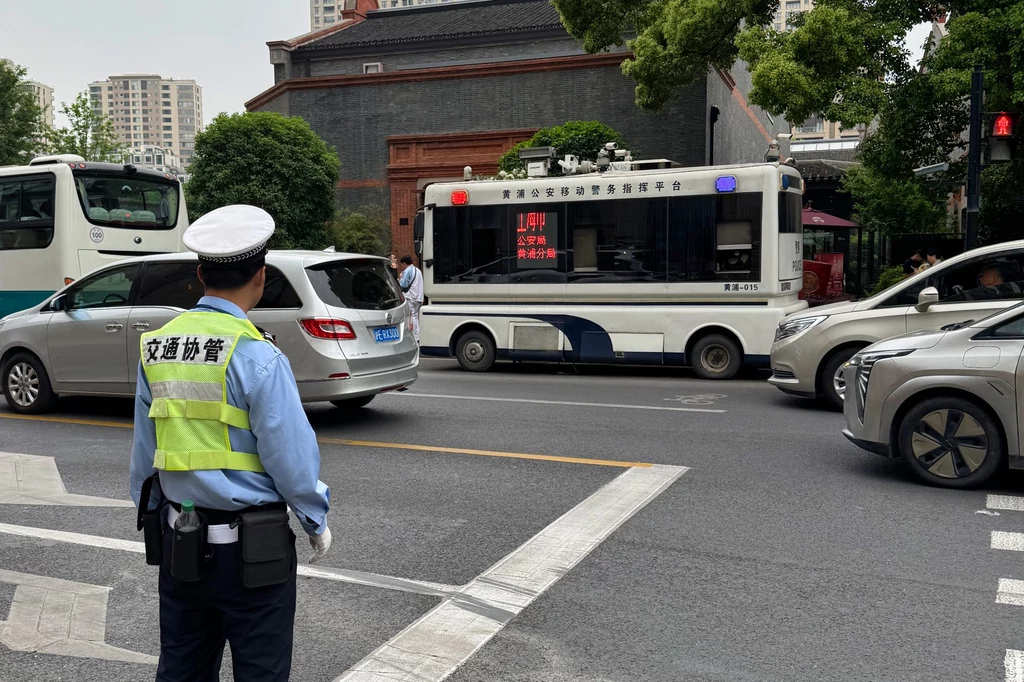 Policjanci kierujący ruchem to bardzo częsty widok – wkraczają, gdy powstaje korek Policjant stojący na ruchliwej ulicy, nadzorując ruch. W tle widoczne samochody, w tym bus policyjny z napisami na ekranie. Otoczenie miejskie, wysoki budynek i drzewa w tle.