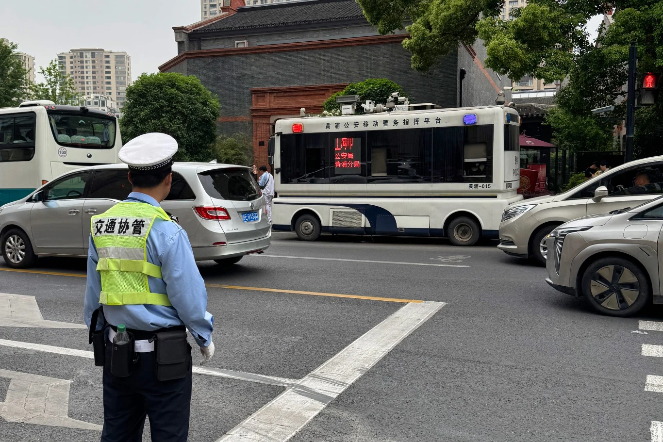 Policjant stojący na ruchliwej ulicy, nadzorując ruch. W tle widoczne samochody, w tym bus policyjny z napisami na ekranie. Otoczenie miejskie, wysoki budynek i drzewa w tle.