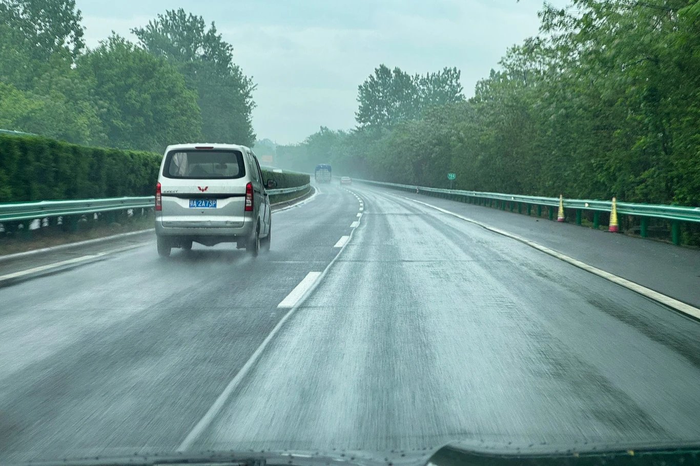 Samochód jedzie mokrą, asfaltową drogą otoczoną zielenią i drzewami, widoczne są barierki po obu stronach drogi.