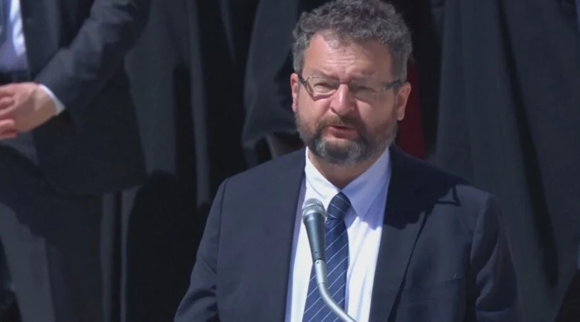 Krzysztof Bronk Mężczyzna w garniturze przemawia na stojąco przy mikrofonie, w tle niewyraźnie widać innych ludzi oraz fragmenty ciemnych ubrań.