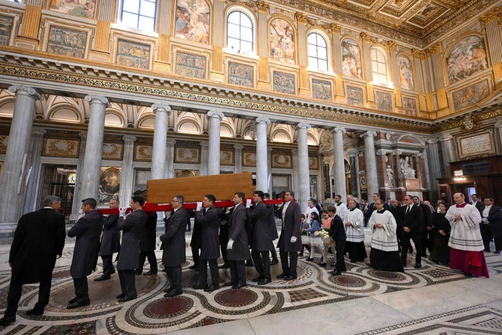 Pogrzeb papieża Franciszka Grupa mężczyzn ubranych na czarno niesie trumnę przez bogato zdobioną salę z kolumnami i freskami na ścianach. Towarzyszą im osoby ubrane w religijne szaty. W tle widać zdobiony sufit i dekoracyjne elementy architektoniczne.