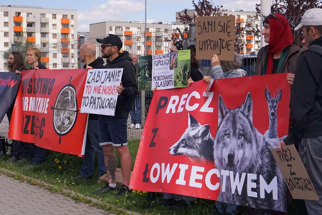 Grupa protestujących trzyma transparenty przeciwko łowiectwu i polowaniom, na których widnieją wilk i lis. Uczestnicy stoją wzdłuż ulicy na tle bloków mieszkalnych. Hasła na plakatach są wyraźnie czytelne, wyrażając sprzeciw wobec polowań.
