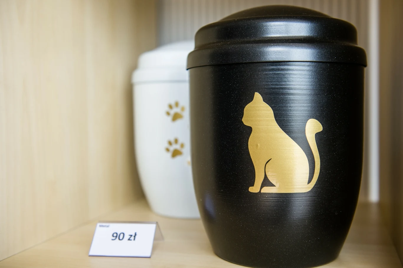 Czarna urna z wizerunkiem złotego kota obok białej urny z motywem łapek na drewnianej półce, obok znajduje się cena 90 zł.