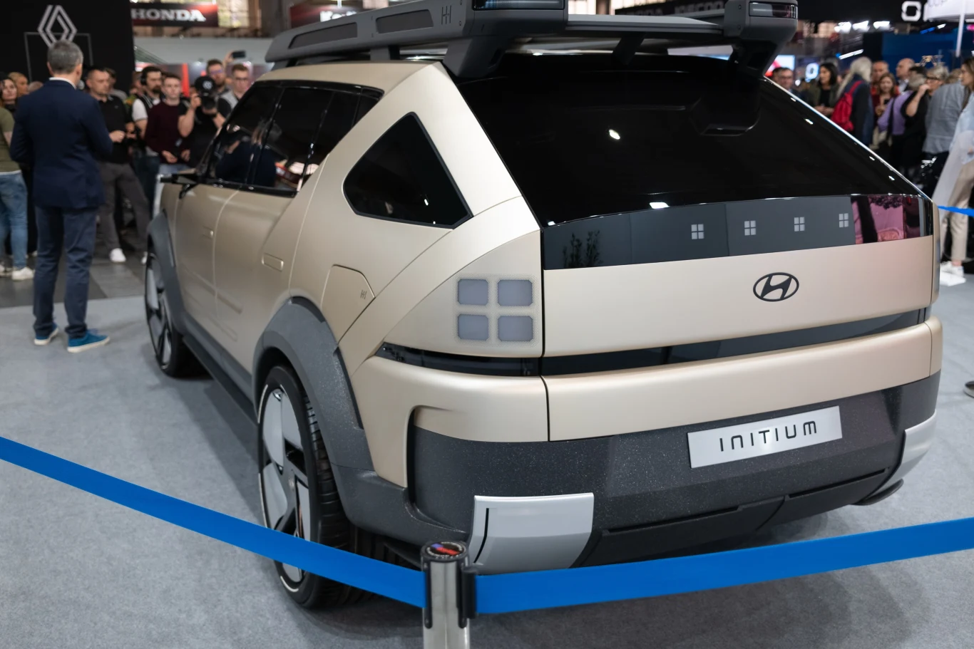 Samochód o nowoczesnym designie z logo Hyundai zaprezentowany na wystawie motoryzacyjnej, otoczony przez osoby oglądające. Samochód w kolorze beżowym z czarnymi elementami oraz oznaczeniem 'Initium'.