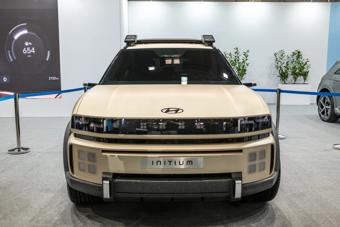 Elegancki samochód Hyundai Initium stoi na wystawie otoczony niebieską taśmą. W tle widać monitury z danymi oraz rośliny ozdobne.