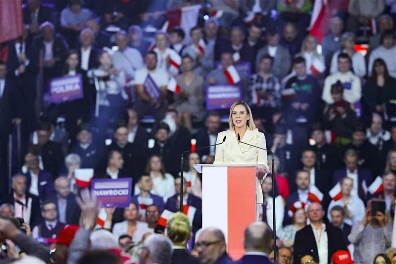 Żona Karola Nawrockiego podczas konwencji w Łodzi Kobieta przemawiająca na tle tłumu ludzi na dużym zgromadzeniu. Widzimy ją na centralnym podium, ubrana w jasny strój, trzyma mikrofon. W tle widać osoby machające flagami Polski oraz trzymające transparenty.