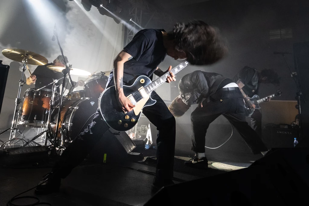 Amenra w akcji Zespół rockowy podczas koncertu na scenie, z dynamicznie poruszającymi się muzykami, w tym gitarzystą wykonującym intensywny riff gitarowy. Dym i światła dodają dramatyzmu występowi.