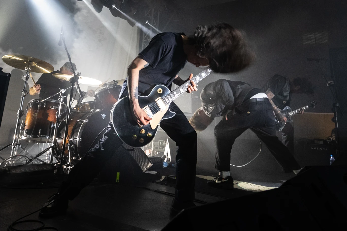 Zespół rockowy podczas koncertu na scenie, z dynamicznie poruszającymi się muzykami, w tym gitarzystą wykonującym intensywny riff gitarowy. Dym i światła dodają dramatyzmu występowi.