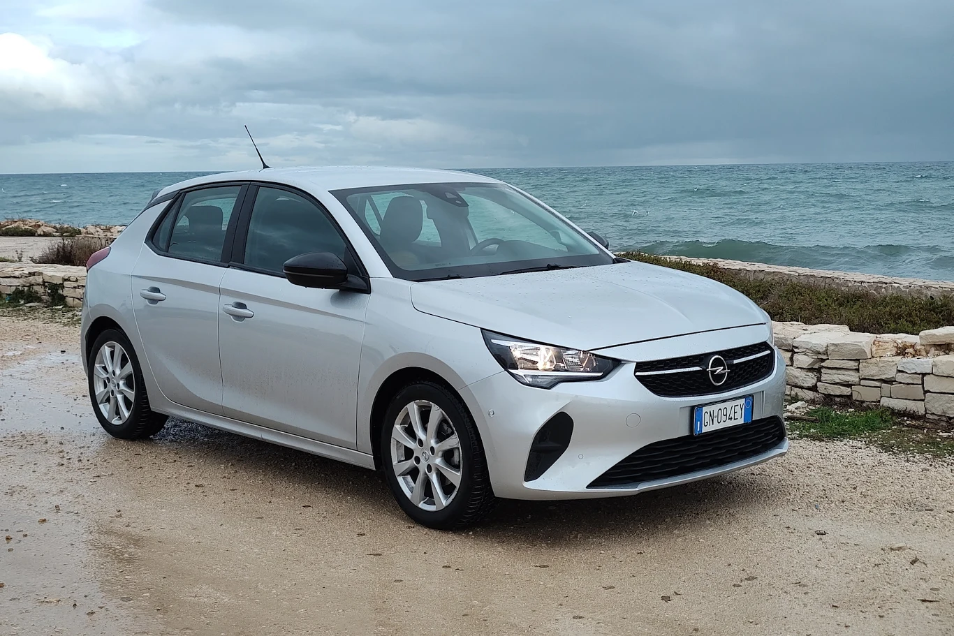Na zdjęciu znajduje się biały samochód marki Opel stojący na tle kamienistego wybrzeża z widokiem na morze. Auto jest zaparkowane na drodze, a w tle widać zachmurzone niebo i zielone krzewy.