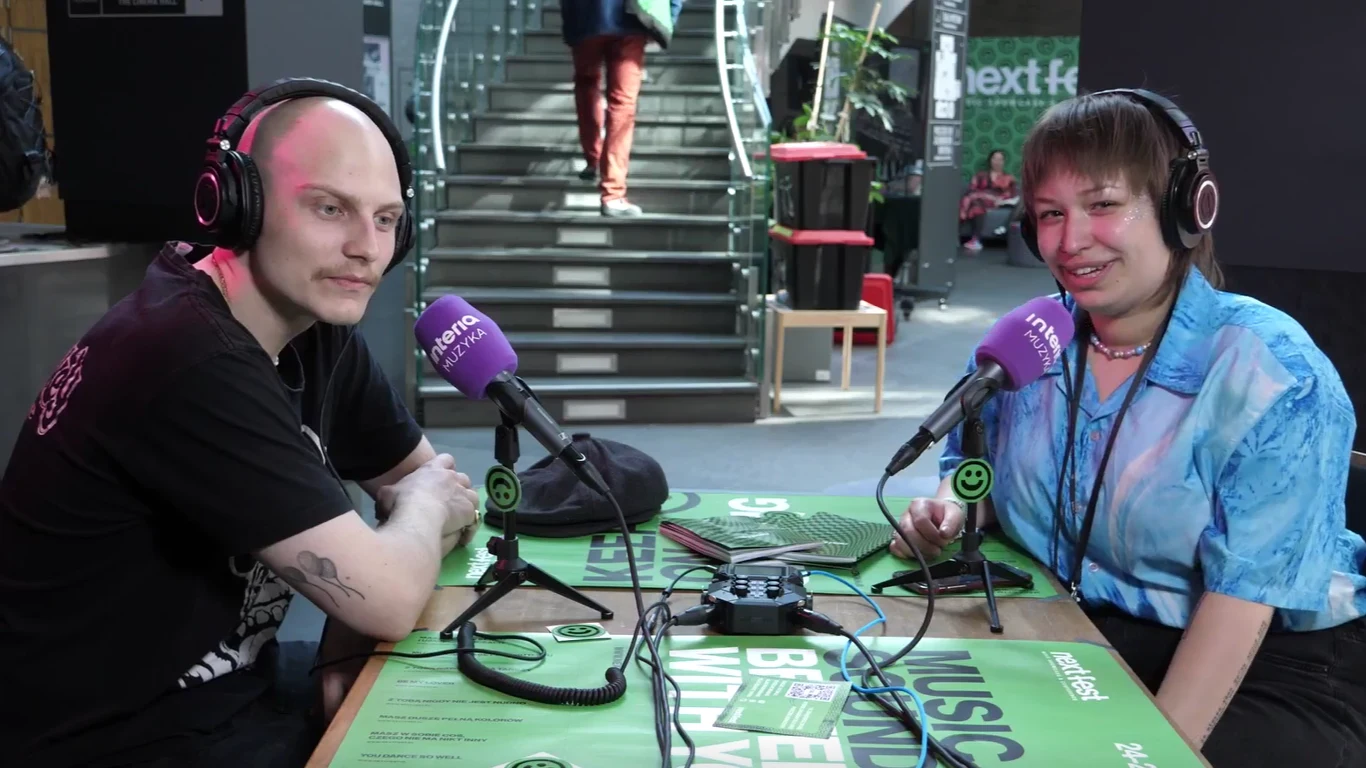 mop wystąpił na tegorocznej edycji Next Festa w Poznaniu Dwie osoby siedzą przy stole, które mają założone słuchawki z mikrofonami. Przed nimi na stole leżą mikrofony i dokumenty. W tle widać schody prowadzące na wyższe piętro oraz wnętrze nowoczesnego budynku.