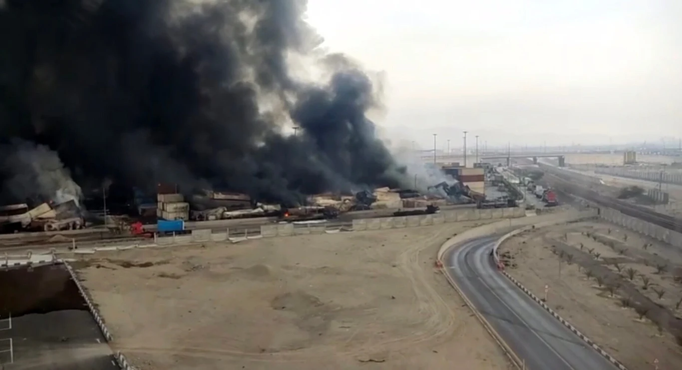 Ogromna eksplozja i pożar w irańskim porcie Czarny dym unosi się z płonących ciężarówek zaparkowanych w przemysłowej części miasta. Widoczna droga biegnie przez opuszczony teren, na horyzoncie widać zabudowania miejskie.