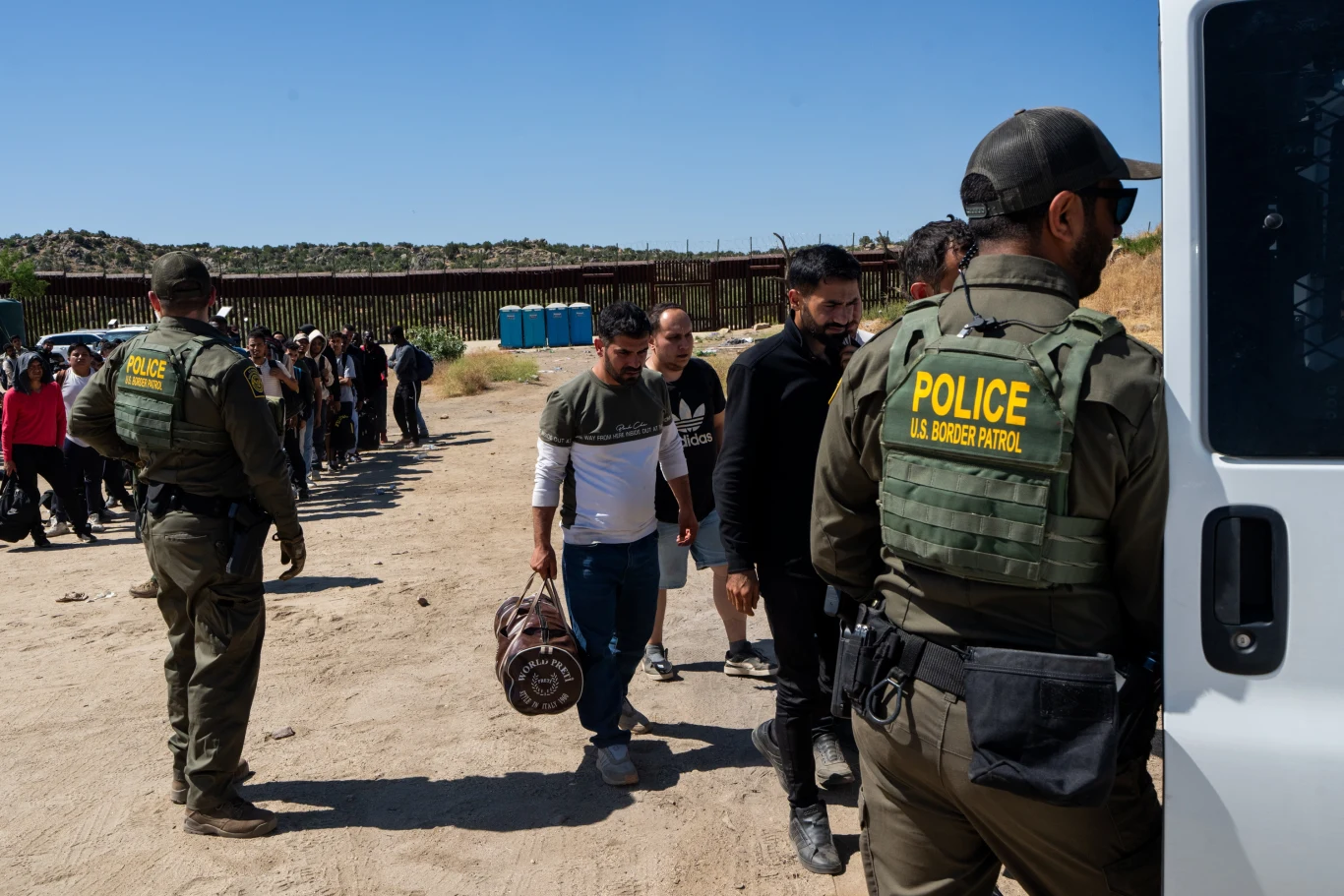 USA. Masowe deportacje. Aresztowano prawie tysiąc migrantów Grupa ludzi zatrzymana przez funkcjonariuszy amerykańskiej straży granicznej w pobliżu granicy, jeden z nich zmierza do pojazdu służbowego. W tle widoczny płot graniczny.