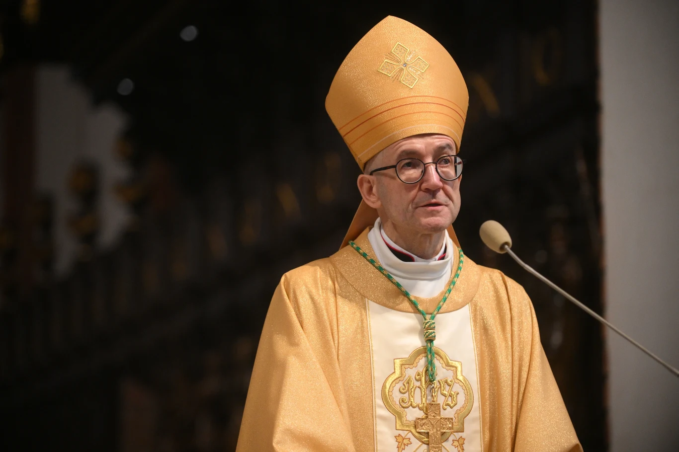 Metropolita warszawski abp Adrian Galbas Biskup w stroju liturgicznym, składającym się z ornatu i mitry, stoi przy mikrofonie w kościele. W tle widoczna ciemność wnętrza świątyni. Jego wyraz twarzy jest spokojny i skupiony.