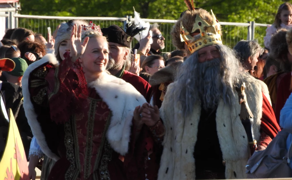Parada historyczna w Warszawie Para ubrana w historyczne stroje, przypominająca postacie królewskie, przechodzi ulicą w trakcie wydarzenia plenerowego. Mężczyzna ma na głowie koronę, a kobieta macha do tłumu, co sugeruje, że są centralną atrakcją imprezy.