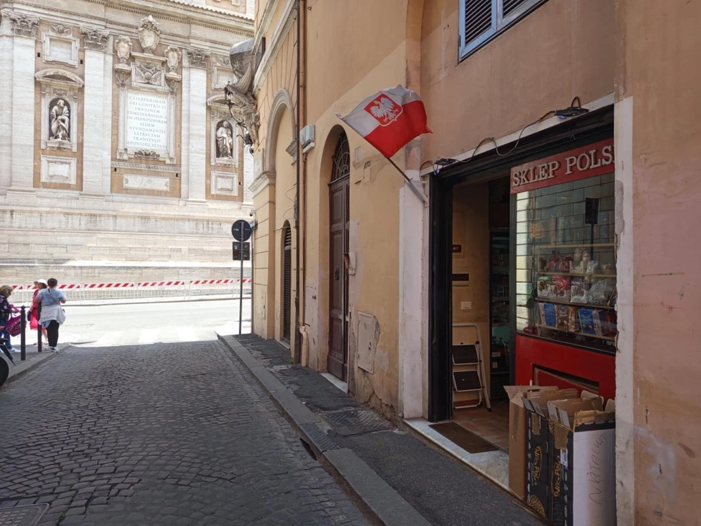 Nad wejściem do sklepu "Karolek" powiewa biało-czerwona flaga, a tuż obok znajduje się Bazylika Matki Bożej Większej Ulica wypełniona historyczną architekturą, po prawej stronie sklep z polskim szyldem i flagą, a z lewej zabytkowy budynek z figurami. Kilka osób spaceruje w tle.