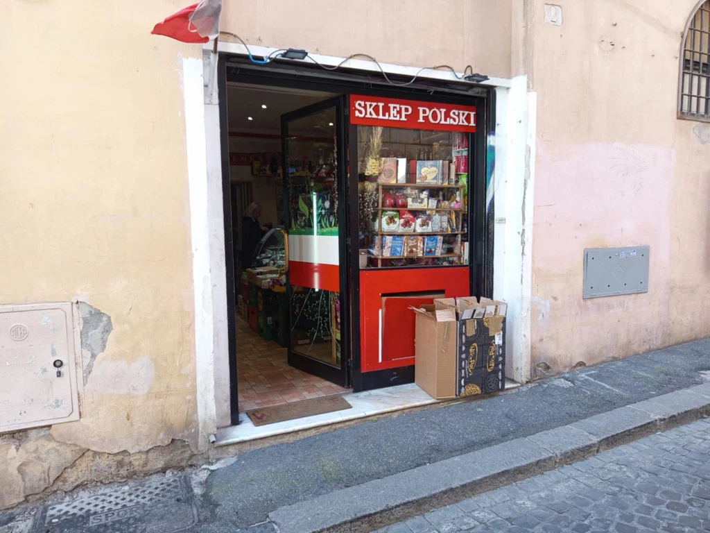 Sklep "Karolek" jest w centrum Rzymu Witryna Sklepu Polskiego z widocznymi produktami na półkach i flagą Polski na drzwiach. Przy wejściu stoją pudełka, a w tle widać wnętrze sklepu.