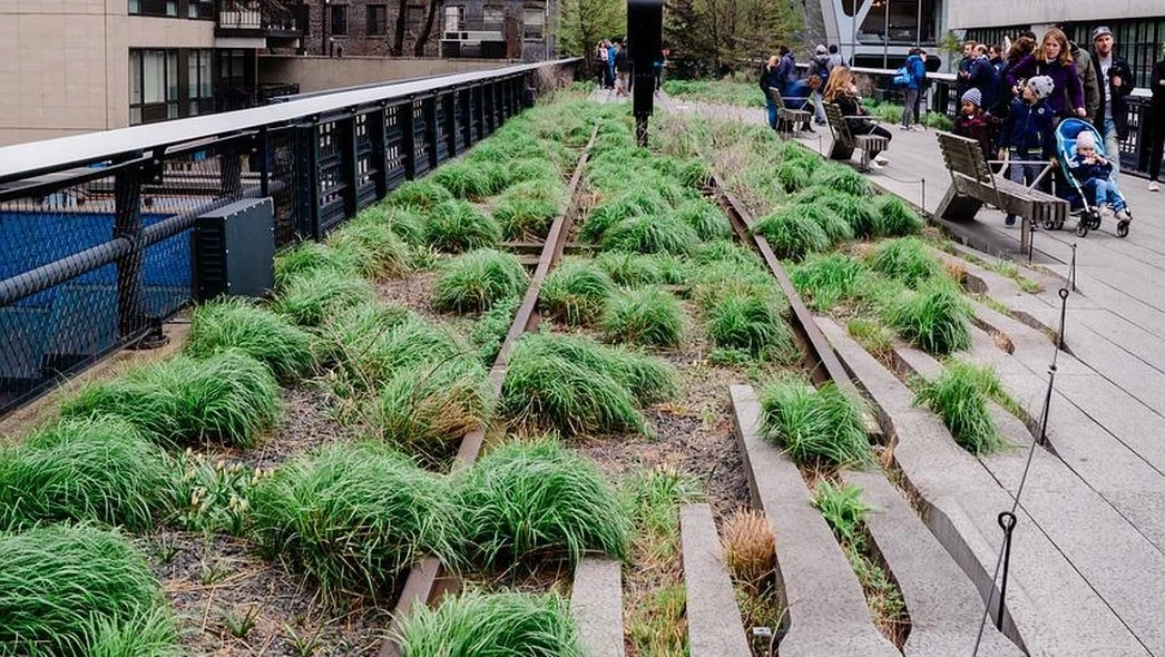 Ogrody miejskie na starych torach kolejowych, z bujną roślinnością i spacerującymi mieszkańcami. W tle kilku ludzi odpoczywających na ławkach.
