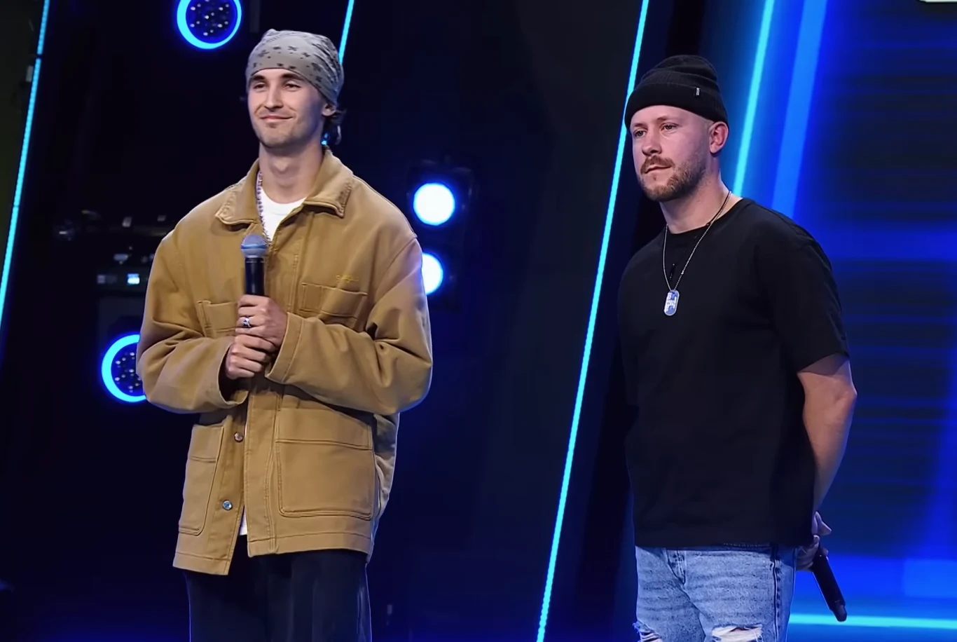 Duet Alien x Majtis w Must Be The Music Dwóch mężczyzn na scenie, jeden trzyma mikrofon. Ubrani są w casualowe stroje, jeden ma na sobie szarą chustę na głowie, drugi czarną czapkę. Jasne tło z niebieskimi światłami sceny w tle.