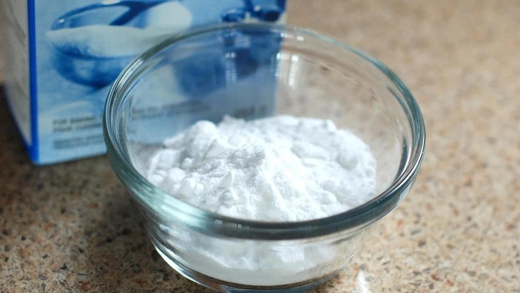 Miseczka wypełniona białym proszkiem, przypominającym sodę oczyszczoną, ustawiona na blacie kuchennym. W tle widać częściowo opakowanie sugerujące zawartość proszku.