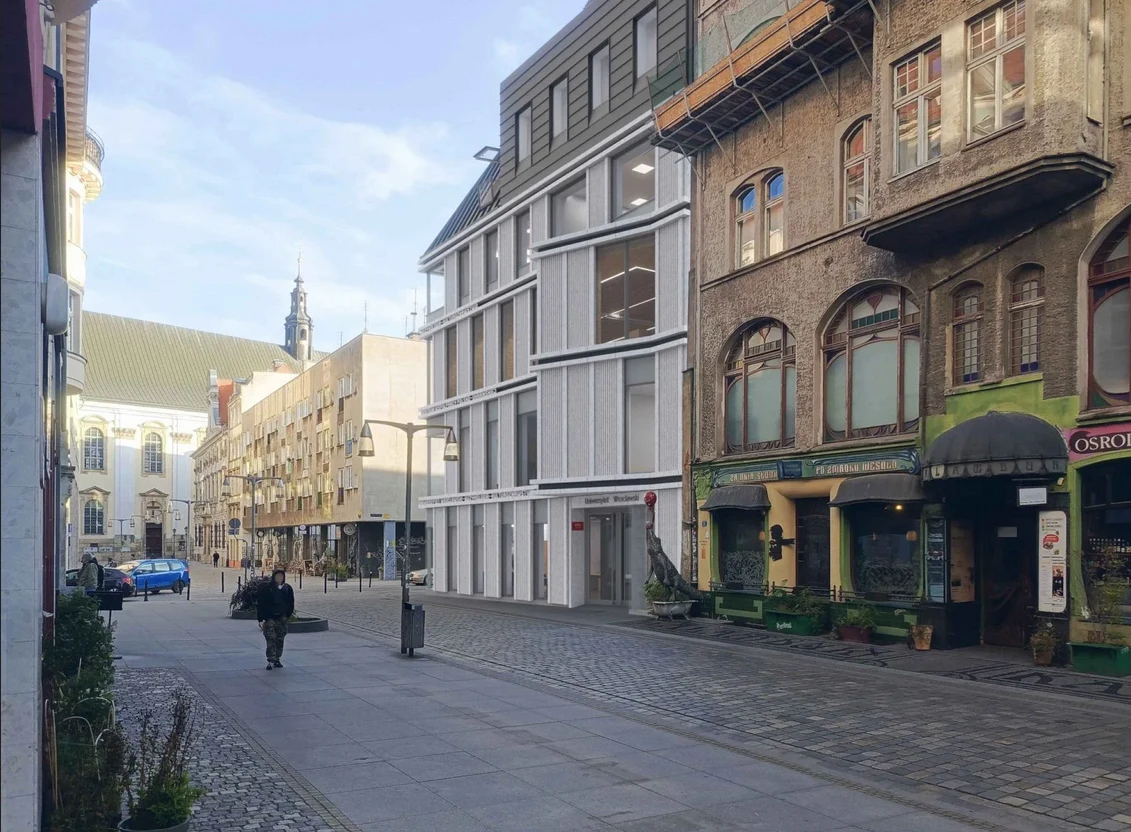 Ulica miasta z zabytkowymi i nowoczesnymi budynkami. Po prawej stronie widać budynki o klasycznej architekturze, zaś po lewej nowoczesna konstrukcja z dużymi oknami. Na drugim planie widać wieżę kościelną, a po ulicy przechadzają się ludzie.